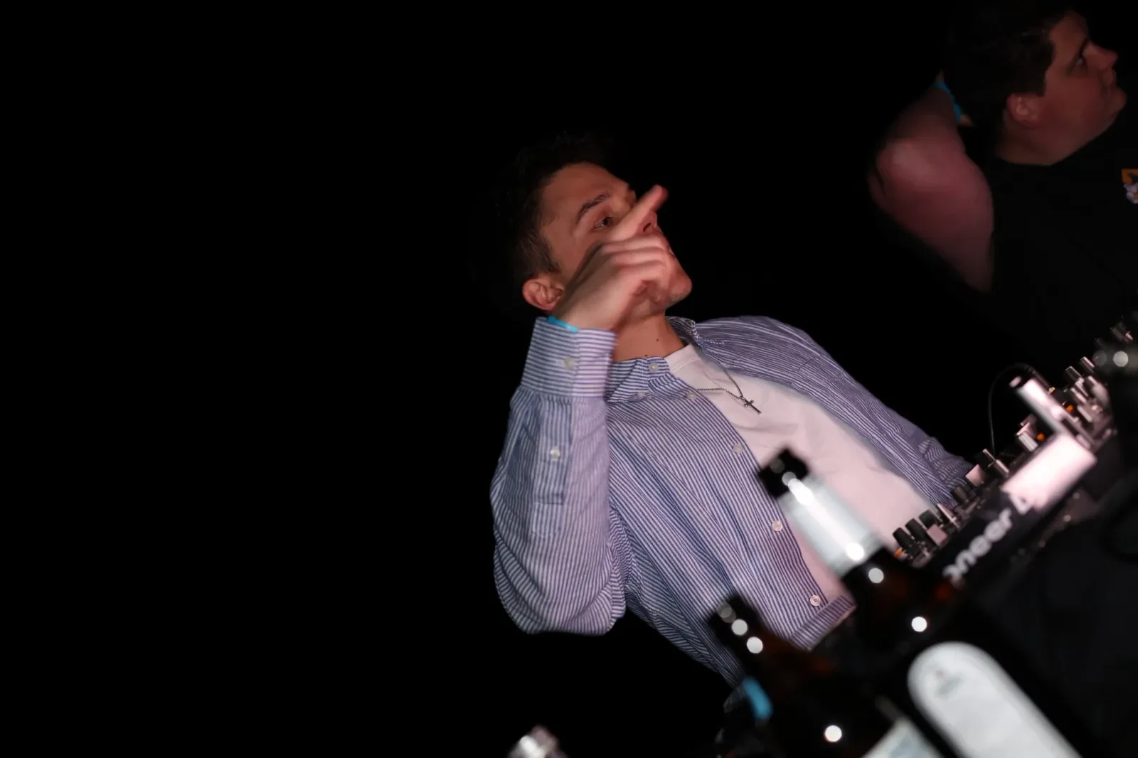 DJ Marian con camisa de rayas azules detrás de la mesa de mezclas, con el dedo índice levantado, entorno oscuro.