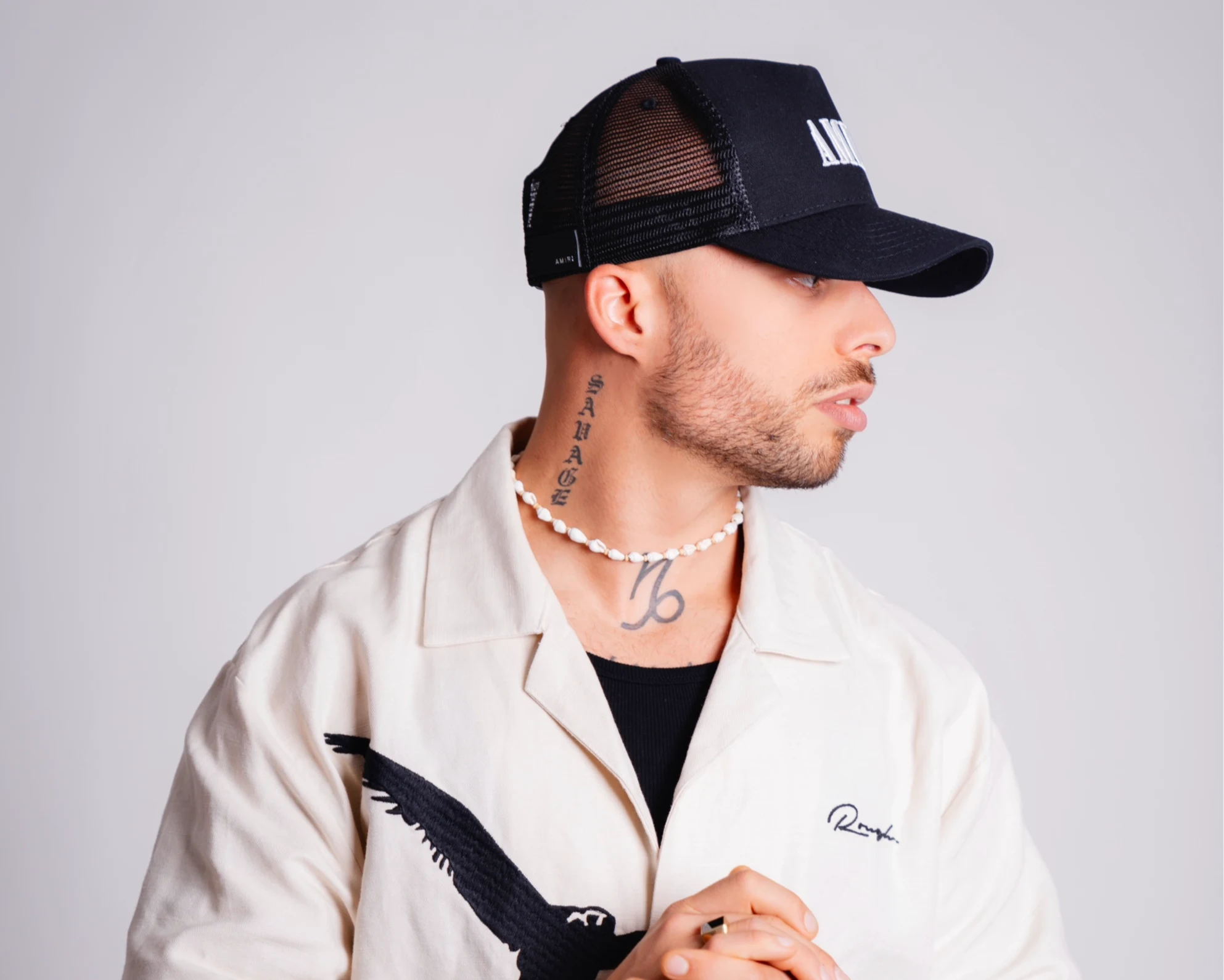 Alex Evans de perfil con gorra negra, camisa color crema, collar de cuentas y tatuaje en el cuello.