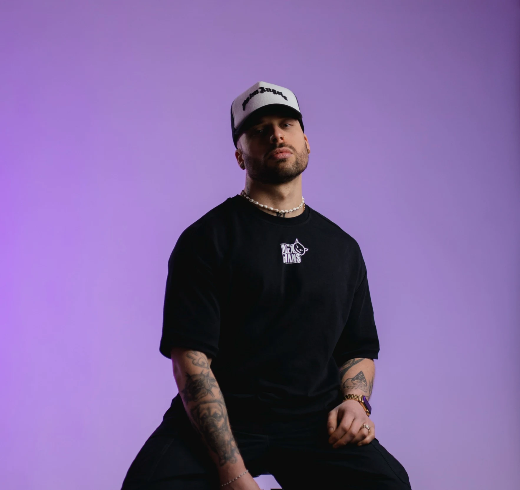 Alex Evans con gorra y camiseta negra oversize ante un fondo de estudio violeta.