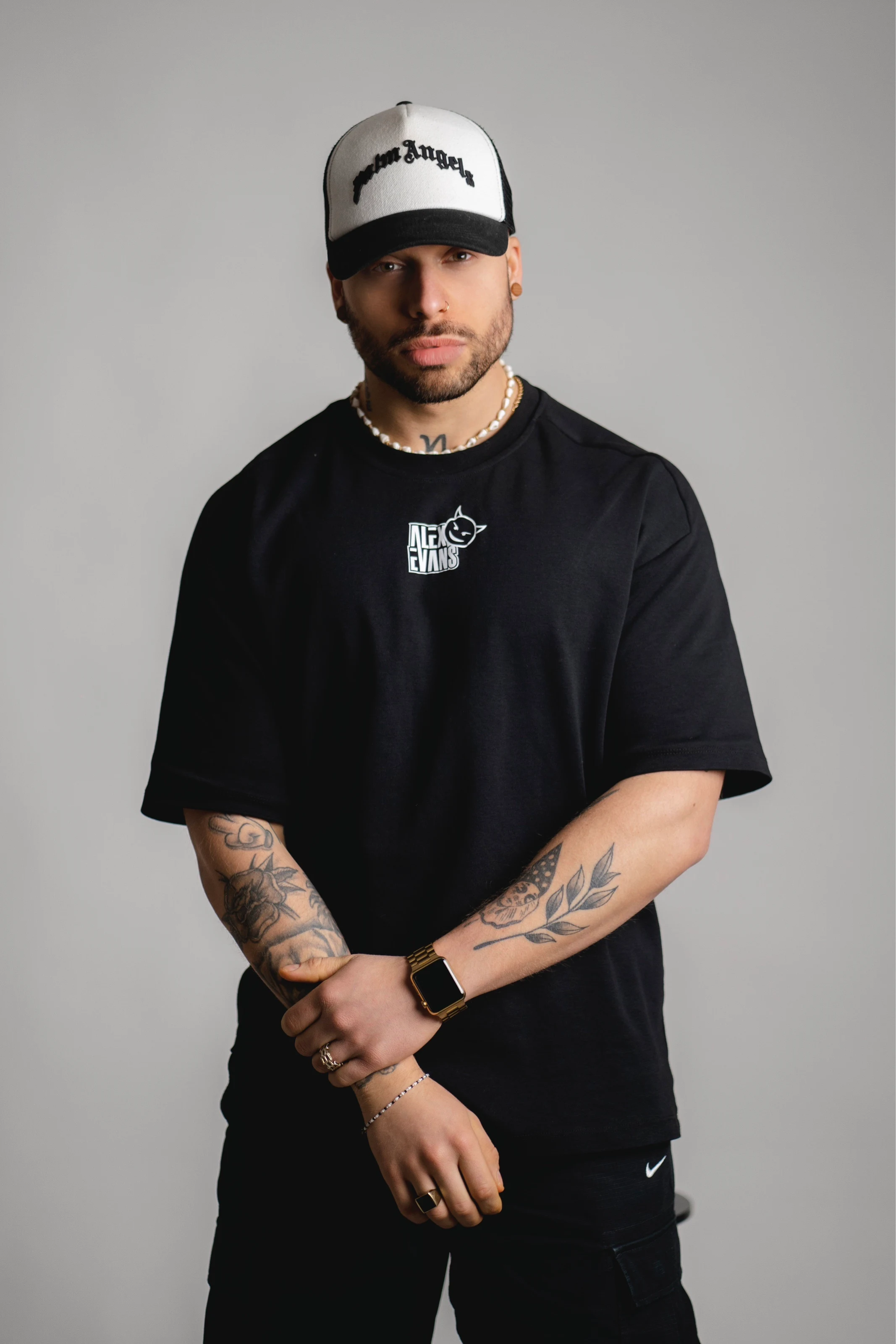 Alex Evans im schwarzen Shirt mit eigenem Logo, weiße Cap, tätowierte Arme vor grauer Wand.